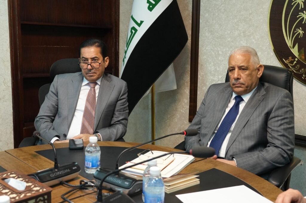691372411_1302136362019759_7137648723887442986_n - مرصد تشريع Two men in gray suits sit at a conference table with microphones, documents, and water bottles nearby.
