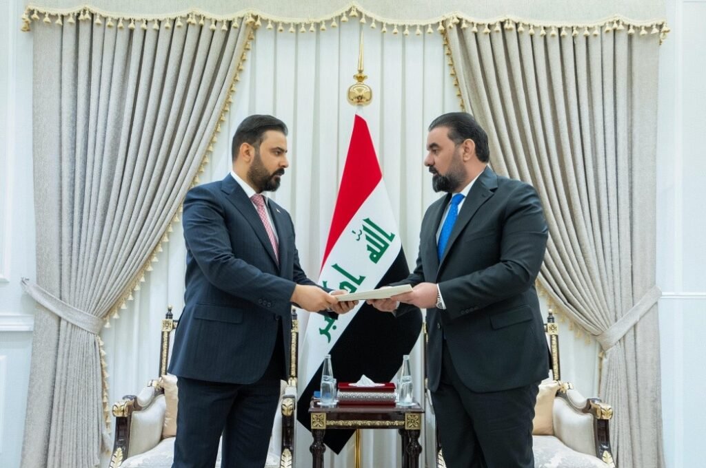 691724654_1299005395666189_6204627845131480096_n - مرصد تشريع Two men in suits exchange a document in a formal room with an Iraqi flag between them on a gold-trimmed backdrop/curtains.
