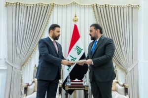 Two men in suits exchange a document in a formal room with an Iraqi flag between them on a gold-trimmed backdrop/curtains.