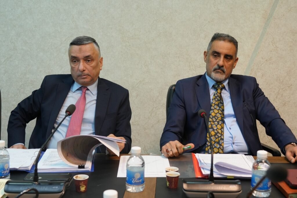693460643_1302136298686432_253705062970477575_n - مرصد تشريع Two men in suits sit at a conference table with microphones, papers, and water bottles, appearing to hold a formal meeting or briefing in progress.